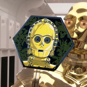 May include: A yellow C-3PO enamel pin with a hexagonal shape. The pin features a detailed depiction of the iconic Star Wars droid against a dark green background with a leaf pattern. The pin is a collectible item.