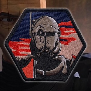 May include: A hexagonal embroidered patch featuring a character from Star Wars. The character is wearing a helmet with a target symbol on the front. The background is a red, white, and blue design.