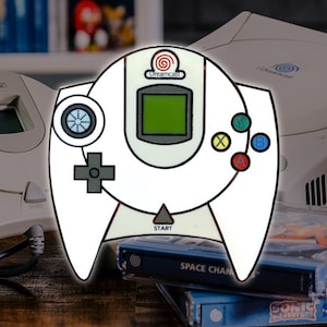 May include: White Dreamcast controller pin with a black and gray directional pad, four colored buttons, and a black and white start button. The pin has a black outline and a white background. The pin is in front of a Dreamcast console and game cartridges.