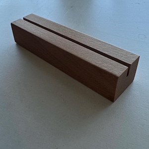 May include: A rectangular wooden block with two parallel grooves cut into the top surface. The block is a warm brown color and appears to be made of solid wood. The grooves are designed to hold cards or signs.
