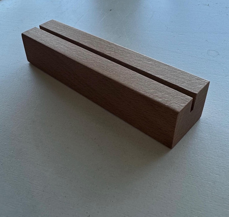 May include: A rectangular wooden block with two parallel grooves cut into the top surface. The block is a warm brown color and appears to be made of solid wood. The grooves are designed to hold cards or signs.