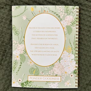 以下が含まれることがあります： 金色の螺旋状の綴じ込みが施された緑と白の花柄のジャーナル。表紙には祈りの言葉と「Ponder Goodness」という言葉が書かれています。