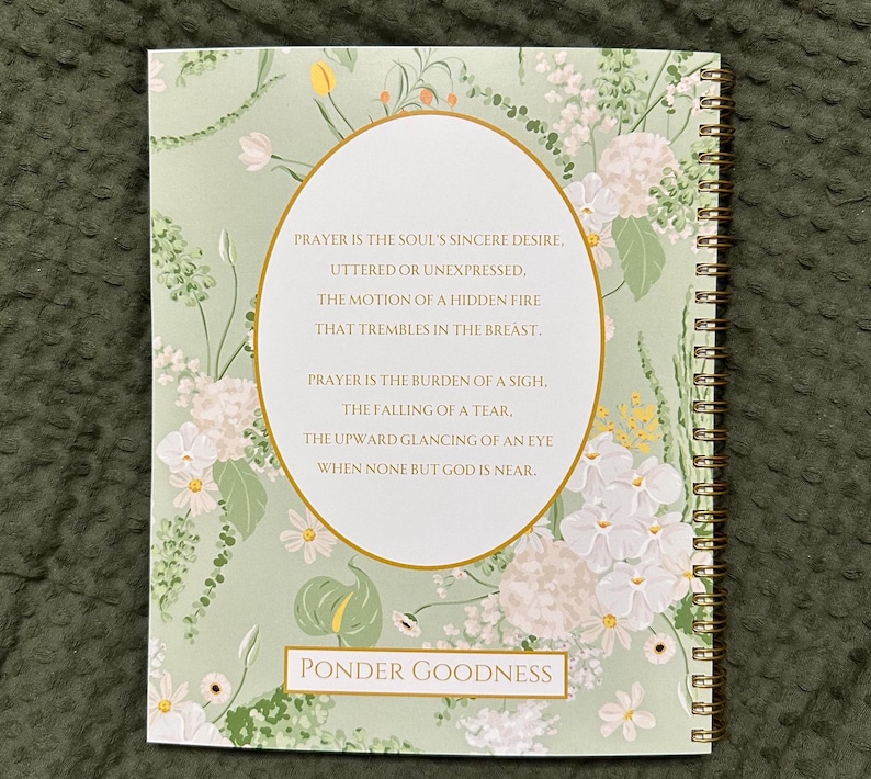 以下が含まれることがあります： 金色の螺旋状の綴じ込みが施された緑と白の花柄のジャーナル。表紙には祈りの言葉と「Ponder Goodness」という言葉が書かれています。