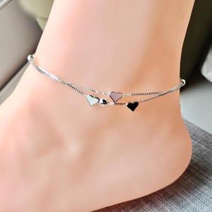 May include: A silver anklet with two delicate chains. The top chain features a small white heart, a silver bead, a pink heart, and a black heart. The anklet is worn on an ankle.