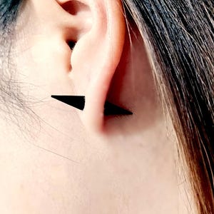 Può includere: Un paio di orecchini triangolari neri indossati sul lobo dell'orecchio di una persona con i capelli scuri.