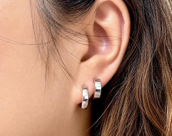 May include: Two small, polished silver-toned hoop earrings worn in the earlobe. The earrings feature a smooth, reflective surface and a simple, circular design that sits close to the skin, creating a clean and understated appearance.