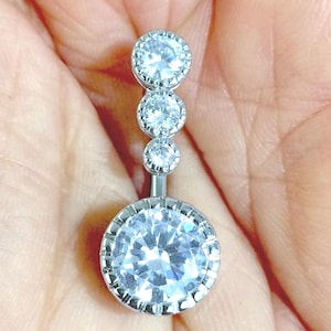 May include: A silver belly button ring featuring four clear, round, sparkling stones. The largest stone is at the base, with three smaller stones above. The ring is held in a hand.