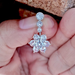 Könnte beinhalten: Ein silberner Bauchnabelring mit floralem Design. Der Ring hat eine Reihe klarer, runder Edelsteine, die zu einer blütenförmigen Ansammlung ähnlicher Steine führen. Der Schmuck wird in einer Hand gehalten.