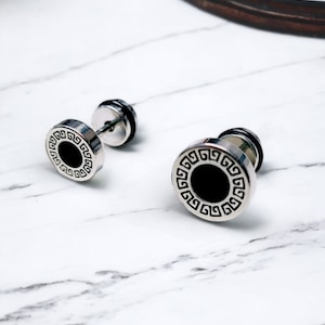 Peut inclure: Une paire de boucles d'oreilles argentées avec des centres noirs et un motif géométrique. Les boucles d'oreilles sont sur une surface en marbre blanc.