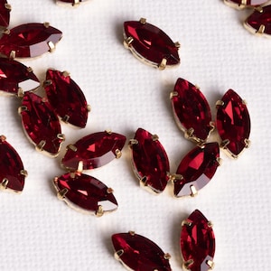 May include: Marquise-shaped red crystal rhinestones with gold-colored metal settings are scattered across a white surface. The crystals have a deep red hue, and the metal settings have a gold finish. These embellishments are suitable for various craft projects.