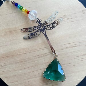 May include: A silver dragonfly suncatcher with a green crystal hanging from it. The dragonfly has intricate cutouts and is attached to a chain with rainbow colored beads.