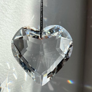 May include: A clear crystal heart-shaped prism hanging from a black string. The prism is faceted and catches the light, creating rainbows.