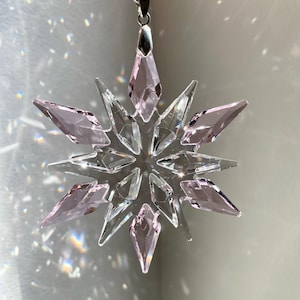 Può includere: Un ornamento a forma di fiocco di neve in cristallo trasparente con un gancio argentato e accenti in cristallo rosa.