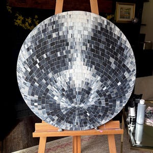 Pu&ograve; includere: Un'opera d'arte circolare con un motivo a mosaico nei toni del grigio e del bianco. L'opera &egrave; esposta su un cavalletto di legno. La superficie &egrave; composta da piccole tessere quadrate disposte in cerchi concentrici, creando un effetto riflettente e strutturato.