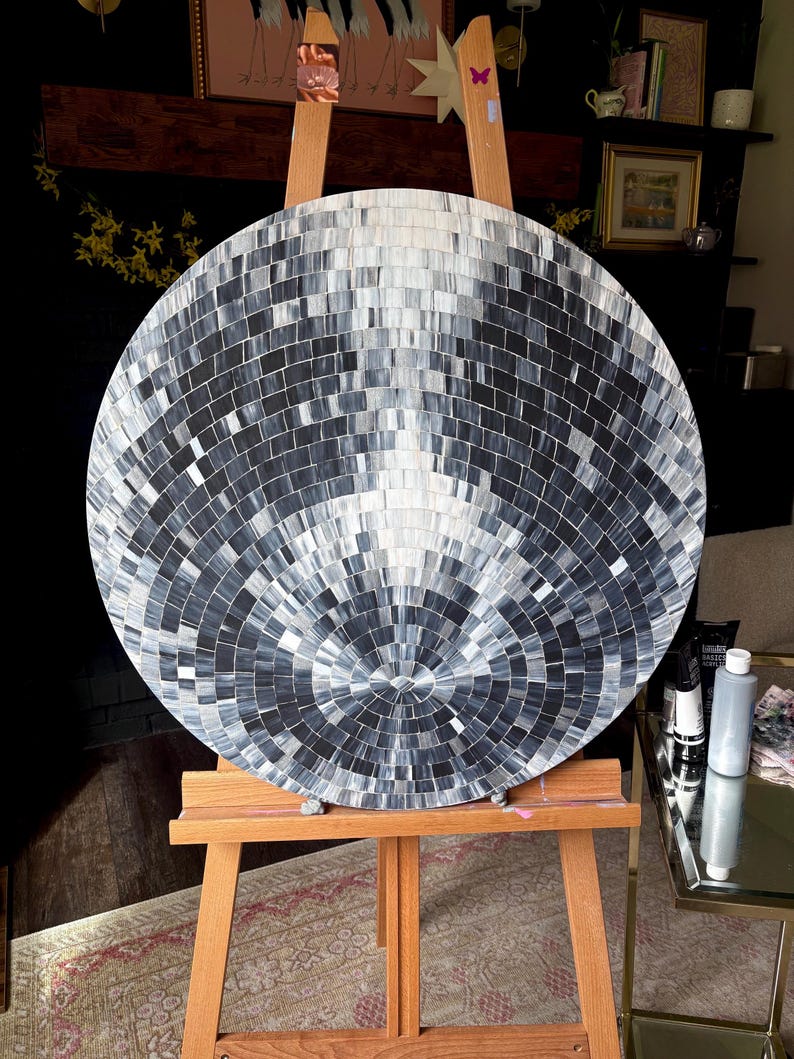 Pu&ograve; includere: Un'opera d'arte circolare con un motivo a mosaico nei toni del grigio e del bianco. L'opera &egrave; esposta su un cavalletto di legno. La superficie &egrave; composta da piccole tessere quadrate disposte in cerchi concentrici, creando un effetto riflettente e strutturato.