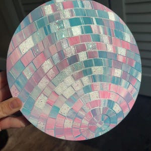 Pu&ograve; includere: Opera d'arte rotonda con un design a mosaico nei colori rosa pastello, blu e bianco. Il mosaico &egrave; composto da piccole tessere quadrate disposte in cerchi concentrici, creando un effetto palla da discoteca. L'opera &egrave; tenuta da una mano.