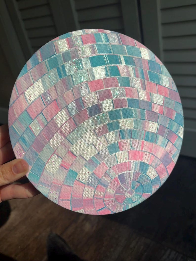 Pu&ograve; includere: Opera d'arte rotonda con un design a mosaico nei colori rosa pastello, blu e bianco. Il mosaico &egrave; composto da piccole tessere quadrate disposte in cerchi concentrici, creando un effetto palla da discoteca. L'opera &egrave; tenuta da una mano.