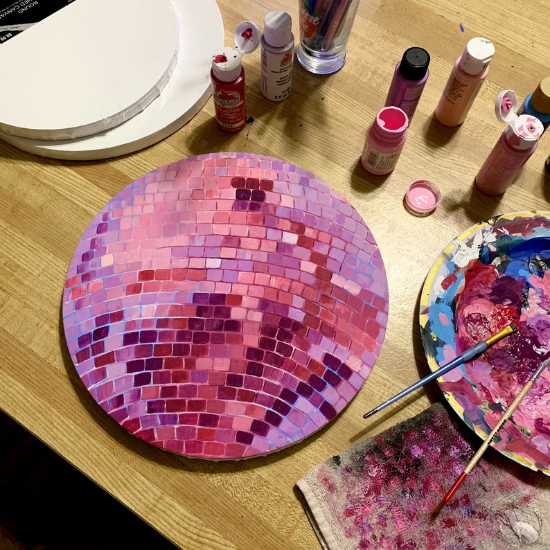 Pu&ograve; includere: Una tela rotonda dipinta per assomigliare a una palla da discoteca, con un motivo a mosaico nei toni del rosa e del viola. Barattoli di vernice e pennelli circondano la tela, suggerendo un progetto artistico in corso.