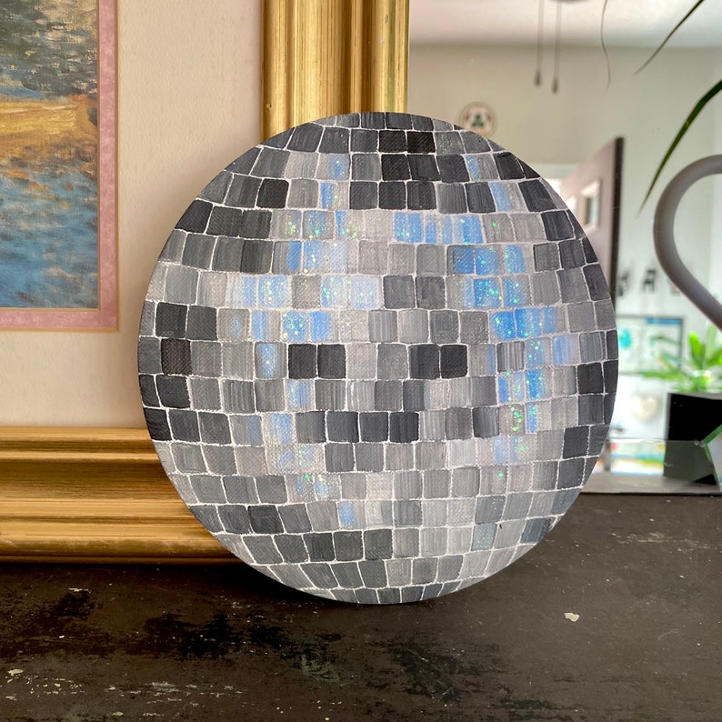 Pu&ograve; includere: Dipinto rotondo di una palla da discoteca, con un mosaico di quadrati grigi, argentati e blu. L'opera d'arte ha un effetto glitterato ed &egrave; posta su una superficie scura. Il diametro &egrave; di circa 20 cm.
