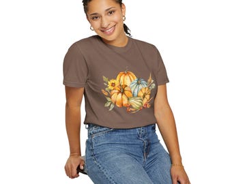 May include: A brown t-shirt featuring a watercolor illustration of pumpkins, sunflowers, and foliage. The design includes orange, yellow, and blue pumpkins, with green leaves and a fall harvest theme. The shirt is paired with blue jeans.