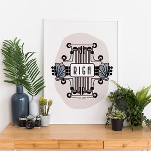 Op de afbeelding: Een witte ingelijste print met een geometrisch ontwerp in zwart, blauw en roze. Het ontwerp bevat het woord "RIGA" en de tekst "Est. 1201". De print wordt weergegeven op een houten bureau met planten en een camera.