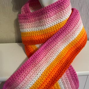 May include: A crochet scarf featuring horizontal stripes in shades of dark pink, light pink, white, and orange. The scarf is wrapped around a white mannequin neck, displaying a textured, yarn-based construction with consistent stitch patterns throughout.