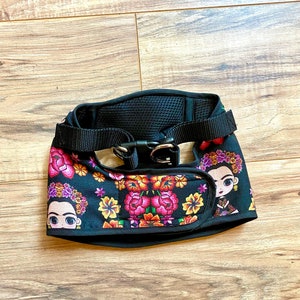 May include: Black dog harness with a floral pattern featuring a cartoon image of a woman with flowers in her hair. The harness has a black adjustable strap and a metal buckle.