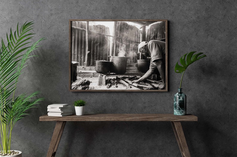 May include: A black and white photograph of a person cooking over a fire in a rustic setting. The person is wearing a hat and is stirring a pot with a wooden spoon. There is smoke rising from the fire.