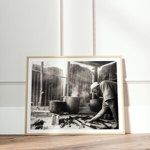 May include: A black and white photograph of a person cooking over a fire in a rustic setting. The person is wearing a hat and is stirring a pot with a wooden spoon. There is smoke rising from the fire.
