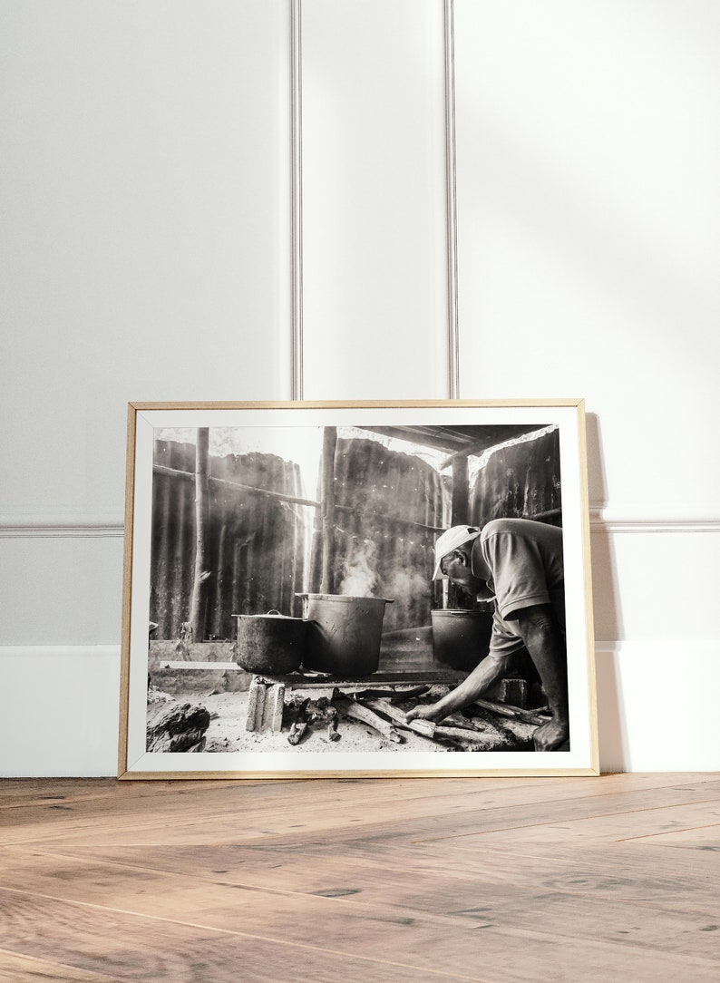 May include: A black and white photograph of a person cooking over a fire in a rustic setting. The person is wearing a hat and is stirring a pot with a wooden spoon. There is smoke rising from the fire.
