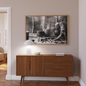May include: A wooden sideboard with two doors and three drawers. The sideboard is a light brown color and has a modern design. A lamp and a bowl are on top of the sideboard. A black and white photograph of a person cooking is hanging on the wall above the sideboard.