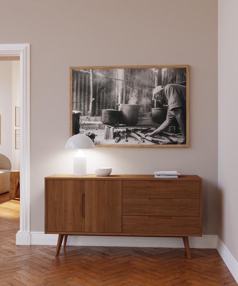 May include: A wooden sideboard with two doors and three drawers. The sideboard is a light brown color and has a modern design. A lamp and a bowl are on top of the sideboard. A black and white photograph of a person cooking is hanging on the wall above the sideboard.