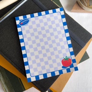 May include: A rectangular notepad with a blue and white checkered pattern and a blue border. The notepad features a red tomato illustration and the text "Jomato". The notepad is on top of a stack of notebooks and a pencil.