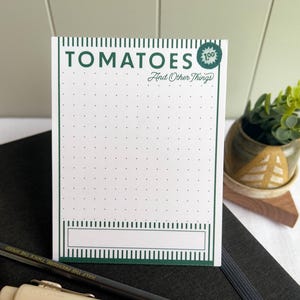 May include: A white notepad with the word "TOMATOES" in green, along with "And Other Things". The notepad has a dotted grid pattern and a rectangular space at the bottom. A pencil and a small potted plant are also visible.