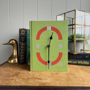 May include: A lime green clock with a unique design featuring red and white semi-circles and square accents. The clock hands are black. The clock is displayed on a vintage table with other decorative items, including books and a brass duck figurine.