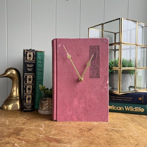 May include: A vintage clock made from a repurposed book with a dusty rose cover. The clock features gold-colored hands and a decorative rectangular design. Other items include a brass duck figurine, books, and a glass and gold terrarium.