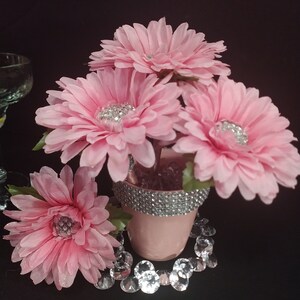 May include: A small pink pot containing four artificial pink gerbera daisies with silver rhinestone centers. The rim of the pot is decorated with a band of silver rhinestones, and clear faceted acrylic gems are scattered around the base.