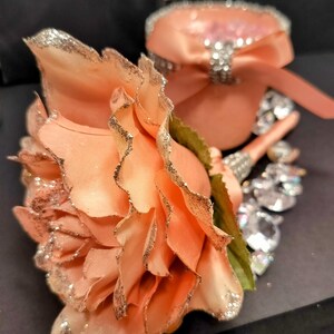 May include: Peach-colored artificial flower with glitter edging, next to a matching ring box with a bow and rhinestone accents. The flower has green leaves and is set against a dark background, suggesting a wedding or special occasion theme.