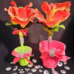 May include: Two artificial rose arrangements. Each rose has red and yellow petals with gold glitter accents. The stems are in decorative pots, one green and one pink, adorned with bows and rhinestones. Decorative crystals are scattered around the base.
