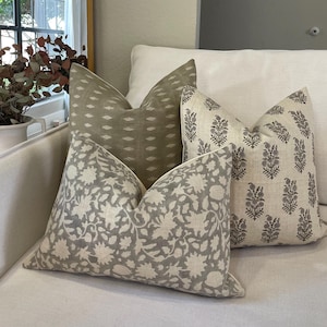 May include: Four decorative throw pillows with floral and geometric patterns in shades of gray and beige rest on a white sofa. The pillows add a touch of elegance to the neutral-toned living space.