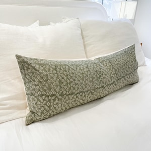May include: A long, rectangular decorative pillow with a sage green background and a white floral pattern. The pillow is placed on a white bed with white pillows, creating a calming and neutral aesthetic. The pillow is approximately 30 inches long.