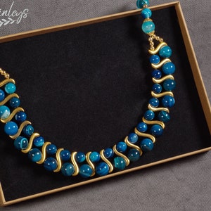 May include: A double-strand necklace made with blue agate beads and gold spacers. The necklace is displayed in a brown box with a black interior.