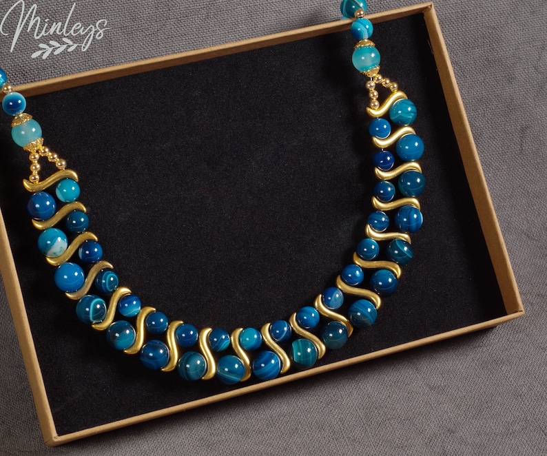 May include: A double-strand necklace made with blue agate beads and gold spacers. The necklace is displayed in a brown box with a black interior.