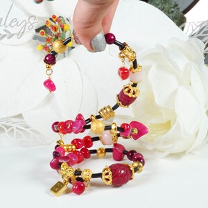May include: A multi-strand bracelet with pink and red gemstone beads, gold accents, and black elastic cord. The bracelet is coiled and features a variety of charms, including a crown and a small gold tag.