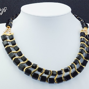 May include: A black and gold necklace with square black beads and gold wire accents. The necklace is on a white mannequin bust.