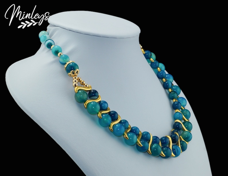 May include: A double-strand necklace with blue agate beads and gold accents. The beads are round and have a swirling pattern. The necklace is on a blue mannequin.