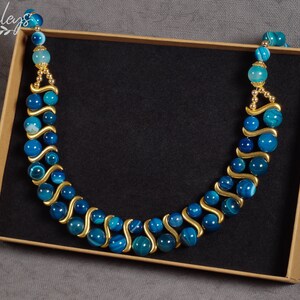 May include: A blue and gold agate beaded necklace in a brown box. The necklace is made of round beads with a wavy pattern and gold accents.