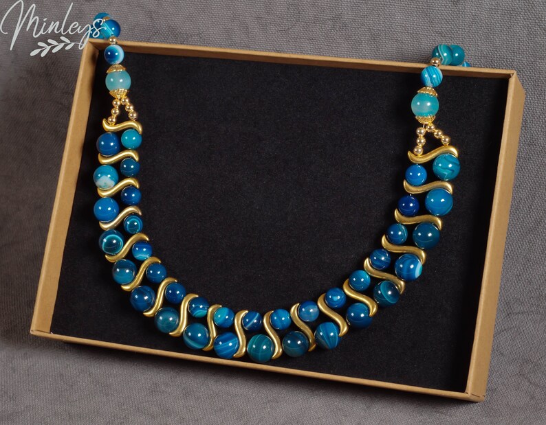 May include: A blue and gold agate beaded necklace in a brown box. The necklace is made of round beads with a wavy pattern and gold accents.