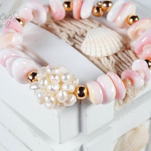 Könnte beinhalten: Ein Armband aus rosa und weißen Muscheln mit goldenen Perlen und einem Perlmutt-Cluster-Anhänger. Das Armband liegt auf einer weißen Holzoberfläche.