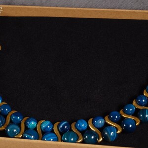 May include: A blue and gold agate beaded necklace with a wavy design. The necklace is displayed in a brown box.